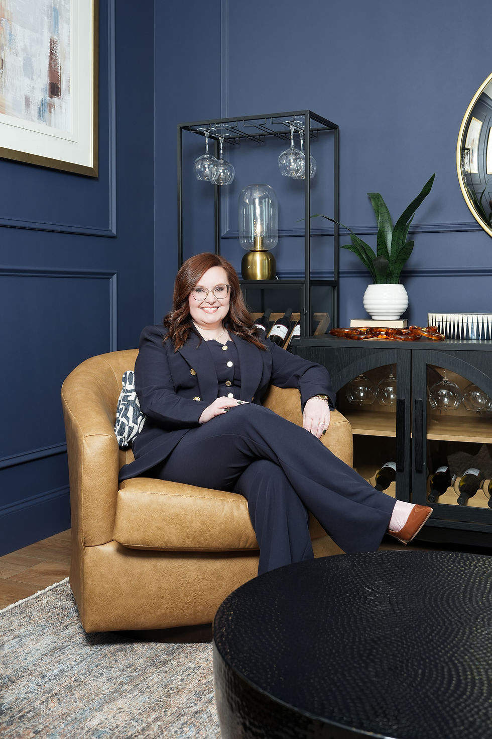 Portrait of Tracy in a stylish interior setting with deep navy, camel, black, and warm neutral tones, vertical/portrait orientation.