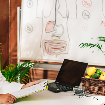 Ernährungsberaterin sitzt am Schreibtisch im Hintergrund ist ein Whiteboard mit der Zeichnung eines menschlichen Verdauungstraktes und ein Obstkorb auf dem Tisch