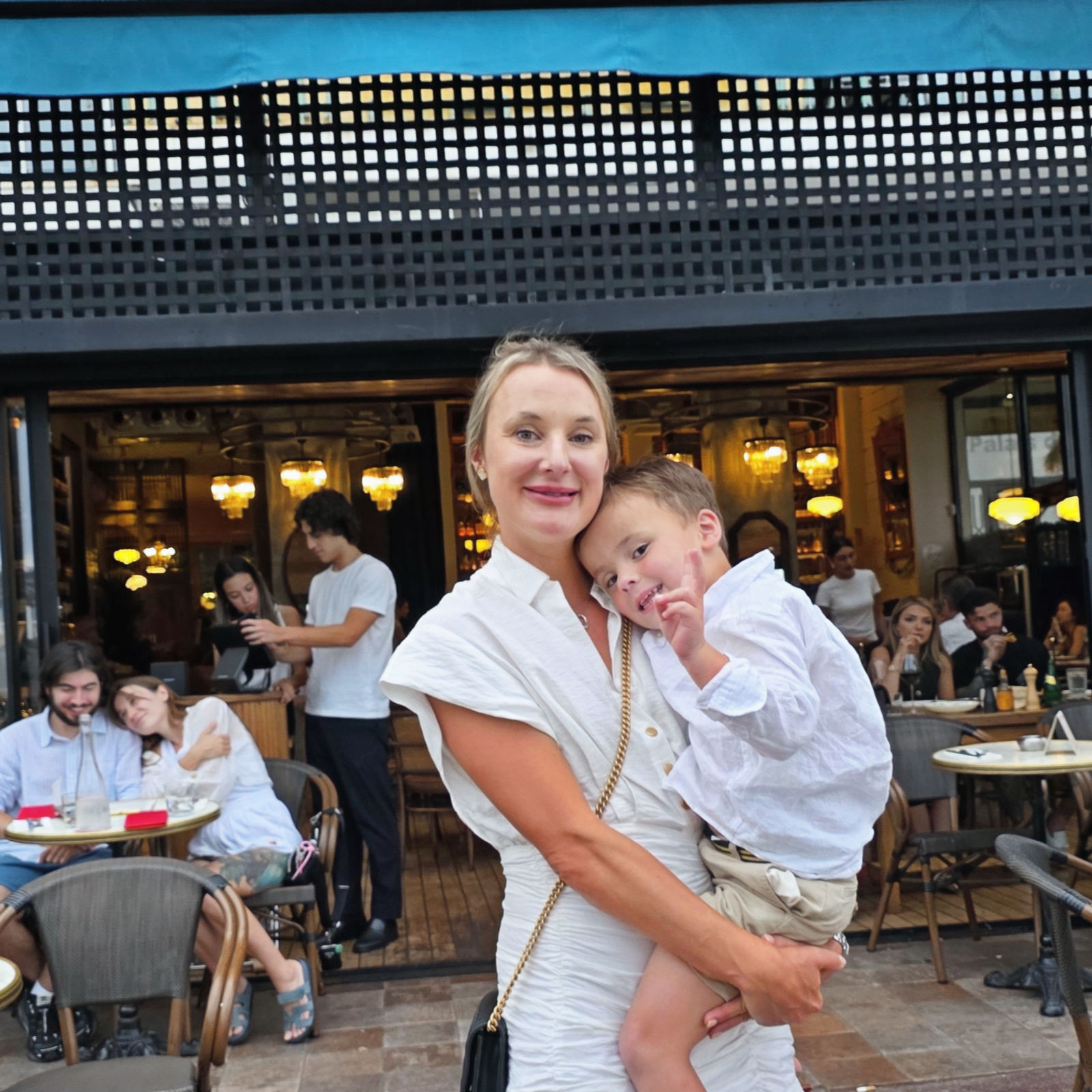 Woman in a white dress holding a child outside a café named "Renée." People dining in the background, warm lights glowing inside.