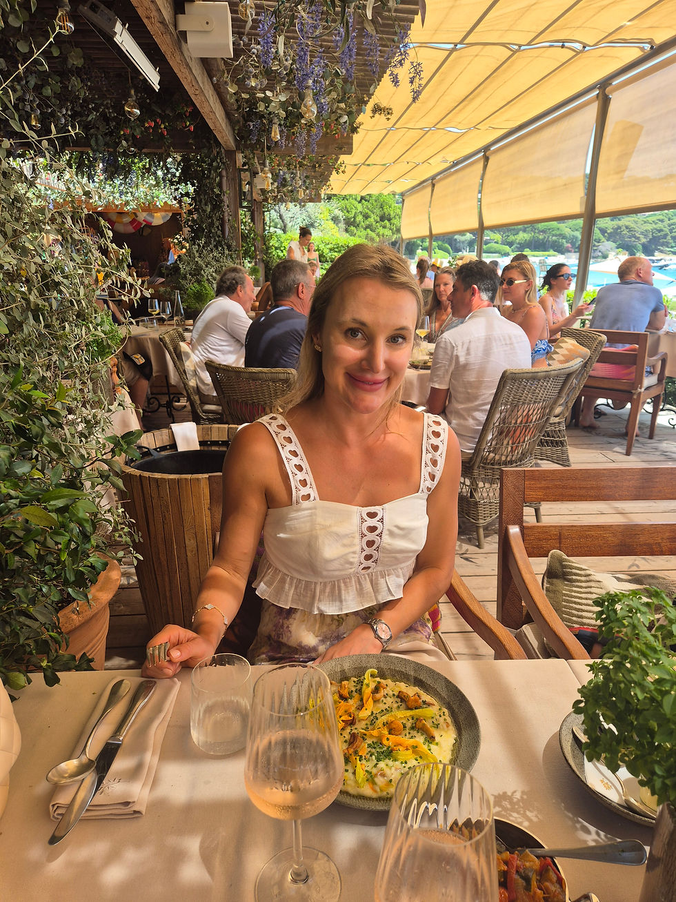 A woman in a white top sits at an outdoor restaurant with a plate of food. She smiles, surrounded by greenery and diners under a canopy.