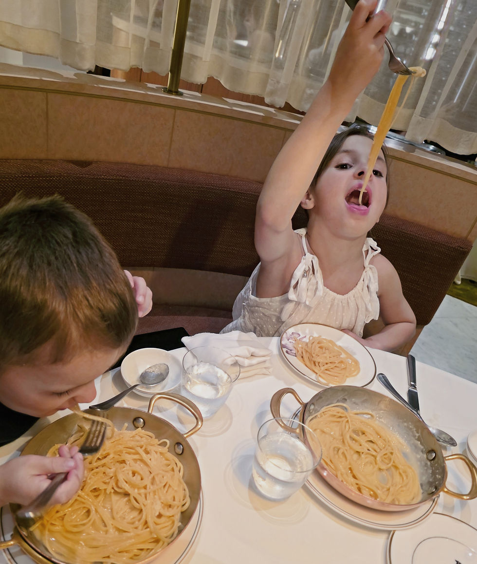 Two kids enjoy spaghetti at a restaurant. One eagerly lifts pasta high, the other leans in to eat. White tablecloth, cozy booth.