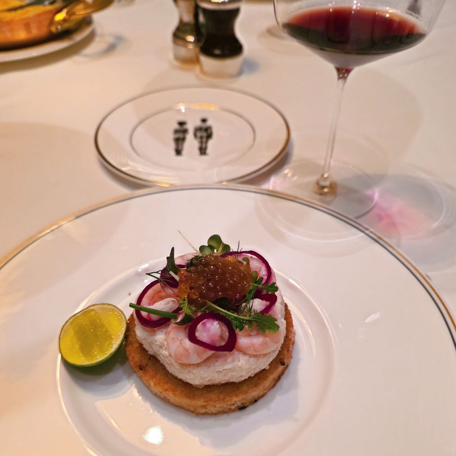 Gourmet appetizer with shrimp, red onion, caviar, and microgreens on a toasted round, lime on side. A glass of red wine in the background.