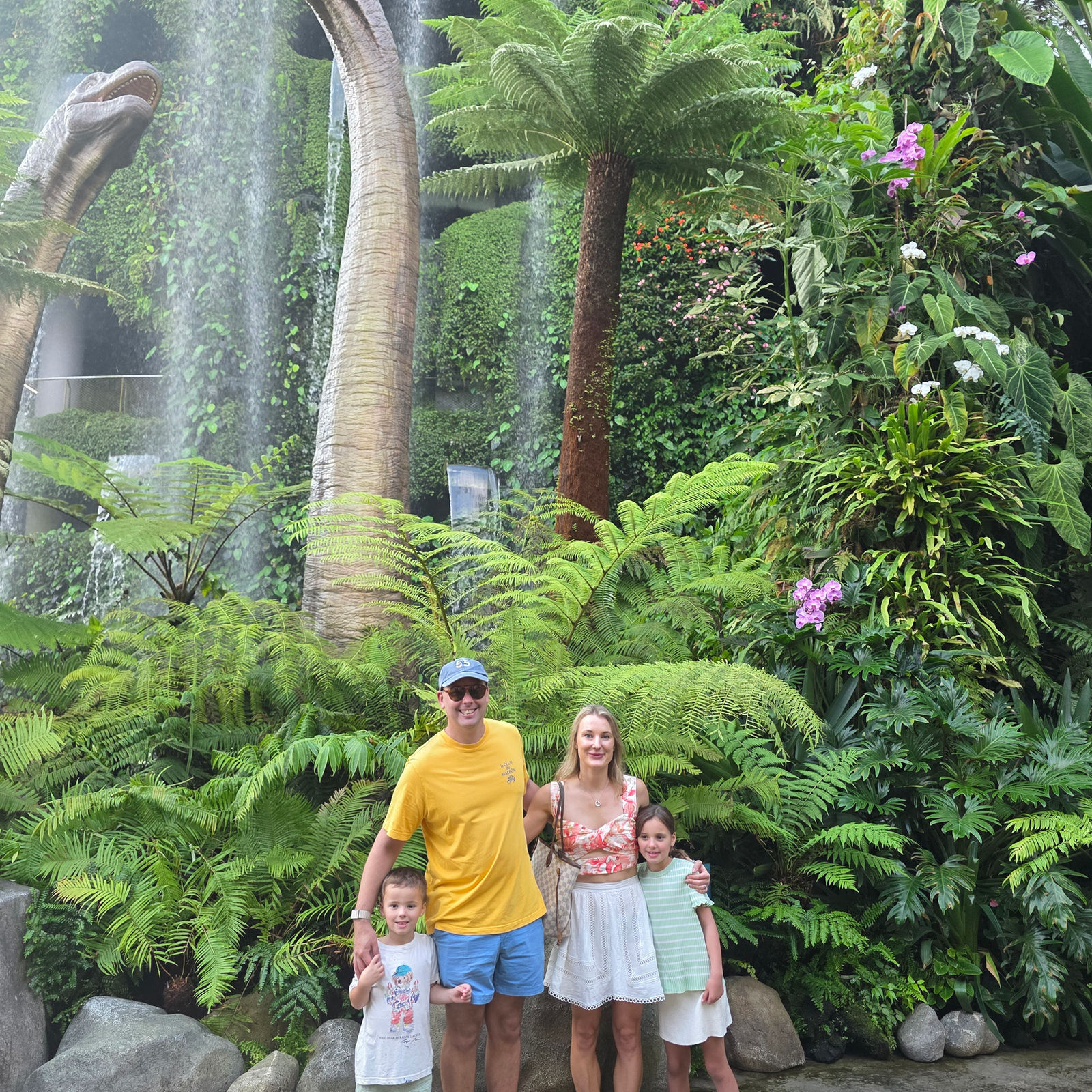 A family of four poses in front of a waterfall and lush greenery with dinosaur statues. The mood is cheerful and tropical.