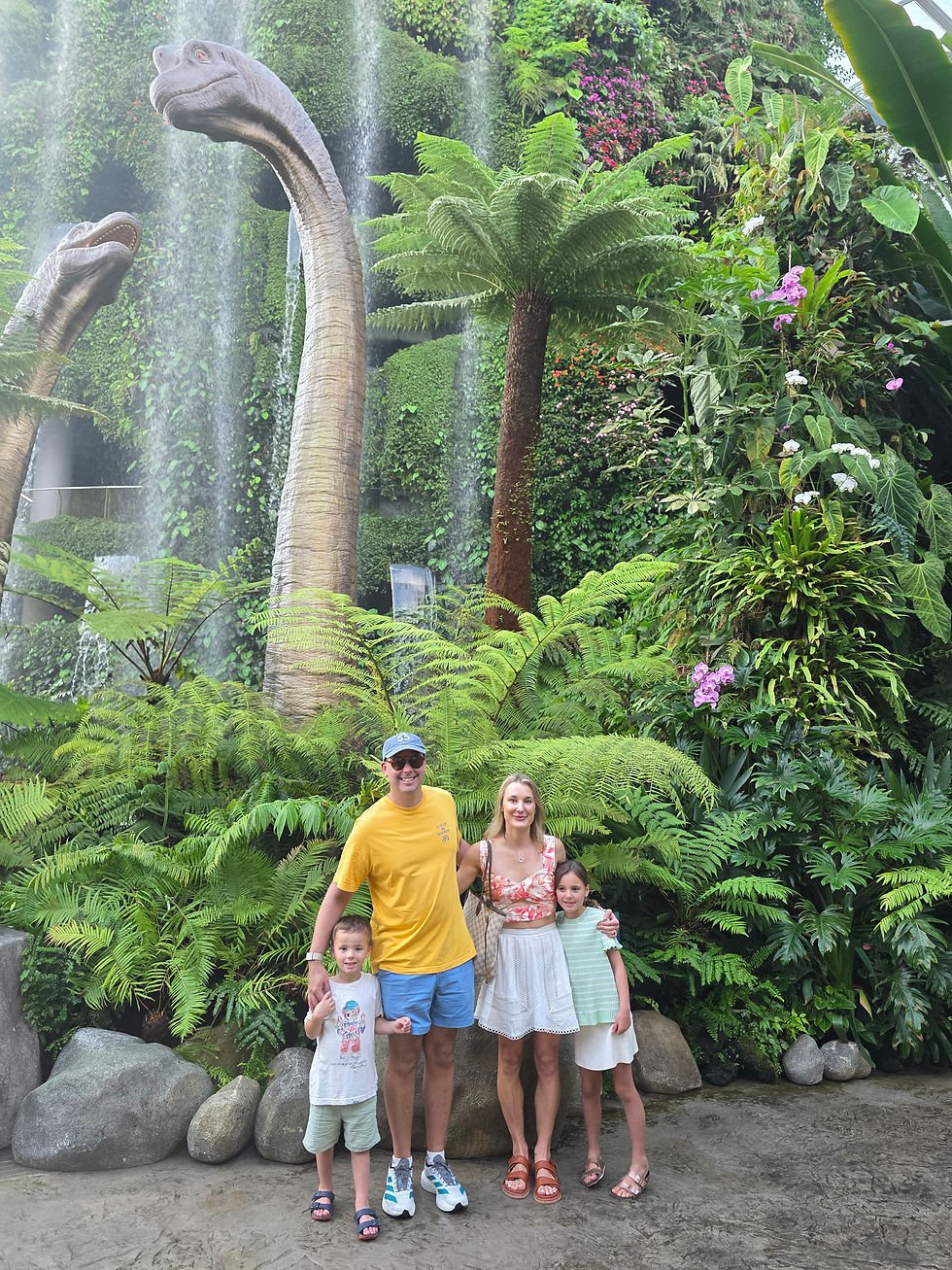 A family of four poses in front of a waterfall and lush greenery with dinosaur statues. The mood is cheerful and tropical.