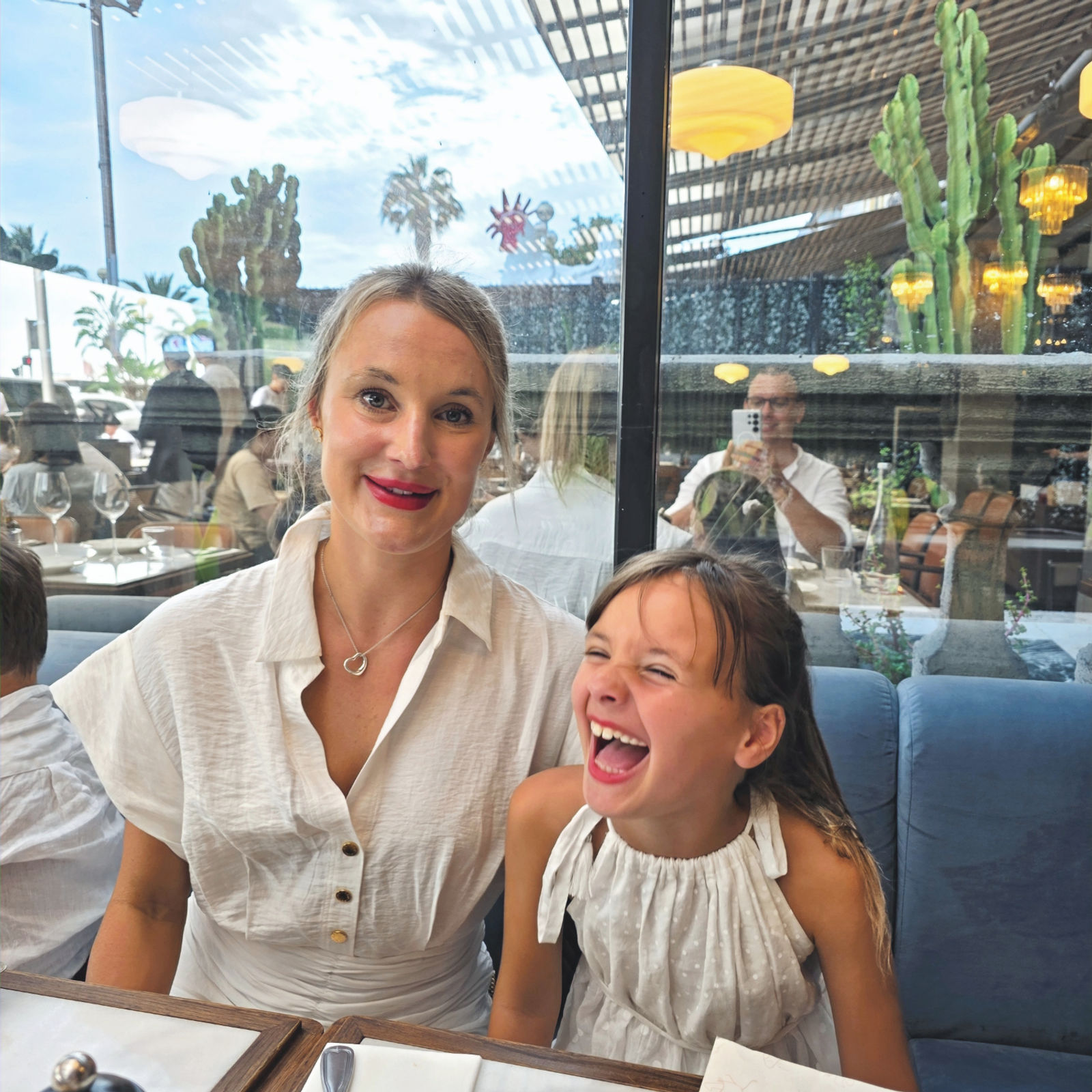 A woman and laughing child in white sit at a restaurant table with menus. Glass window reflects palm trees and blue sky outside.