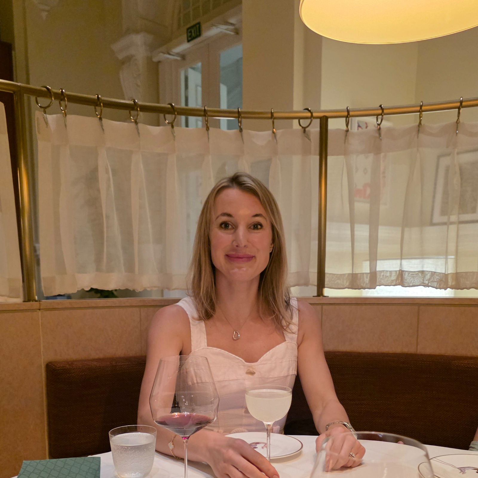 Woman in a white dress sitting at a restaurant table with wine and cocktails. Soft lighting and elegant decor create a warm ambiance.