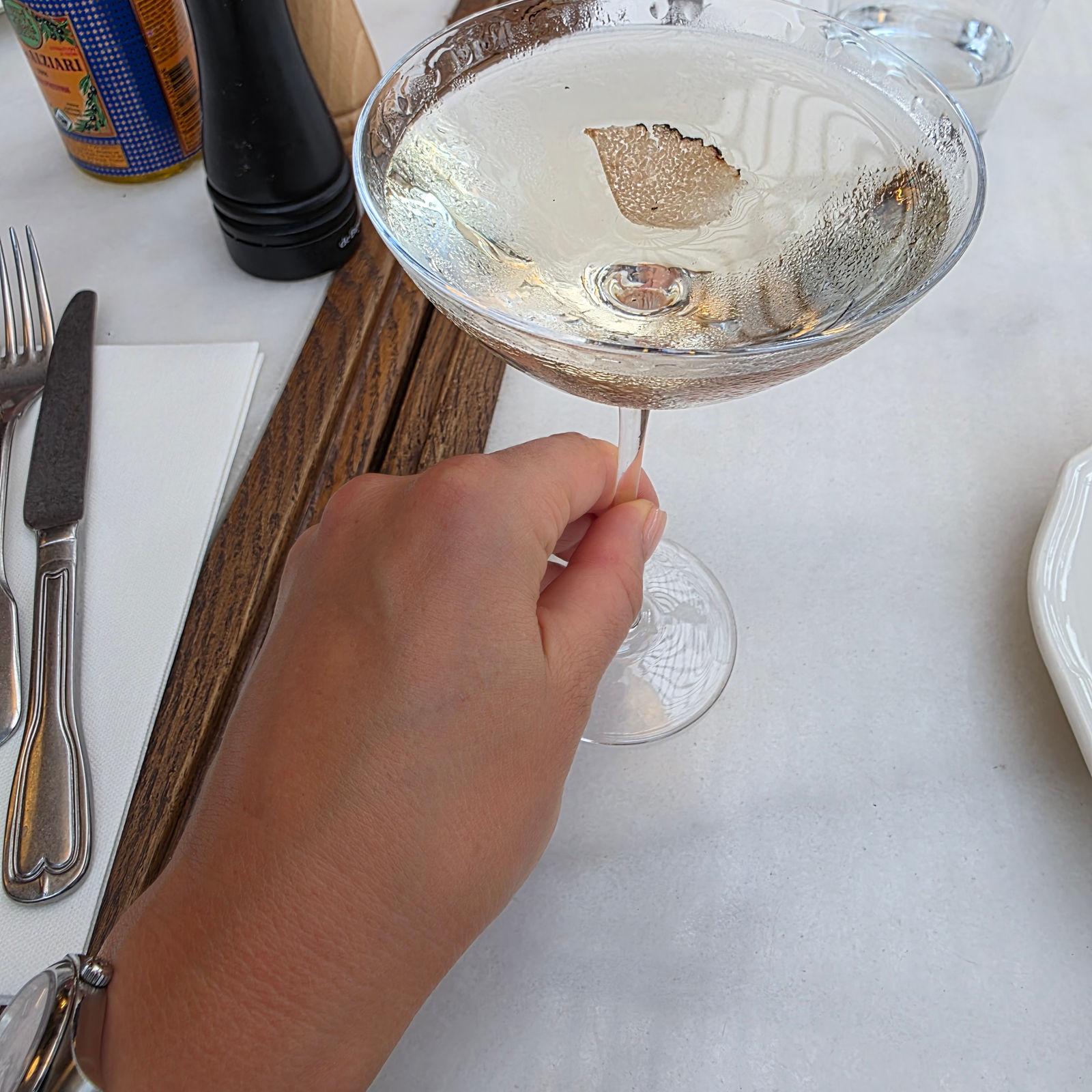 Hand holding a glass with a drink, ice cube, and leaf garnish on a white table. Cutlery, a pepper mill, and a drink bottle are nearby.