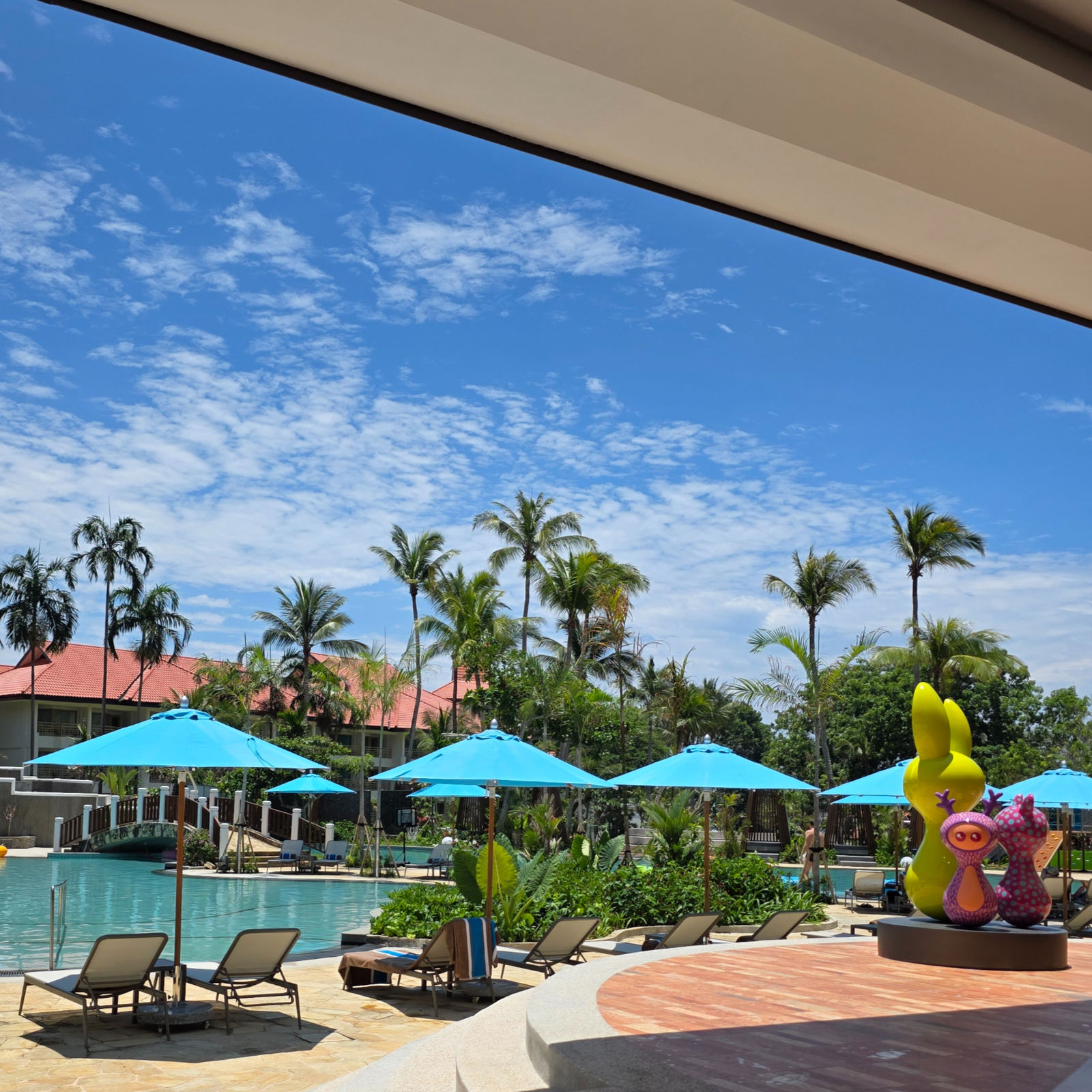 Poolside scene with blue umbrellas, lounge chairs, and colorful sculptures. Palm trees and buildings in background under a clear sky.
