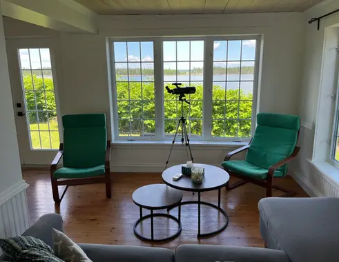 Living room of The Groundswell's Cottage with two green chairs, a couch, coffee table and telescope looking out on the ocean through a big bay window