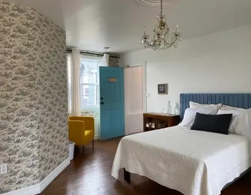 A double bed with a blue headboard in a room with a blue door, yellow chair and one wall papered in wallpaper