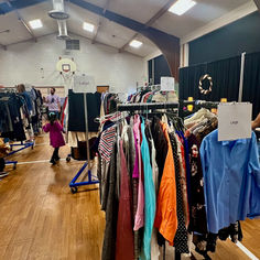 Clothing racks with various sizes are displayed in a gym. A child in pink and adults browse clothes. Signs read "Large" and "Extra Large."