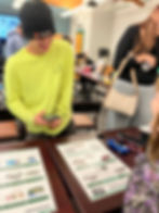 A person in a yellow shirt and black beanie smiles while holding a phone, talking to another person. Toy cars and product posters on the table.