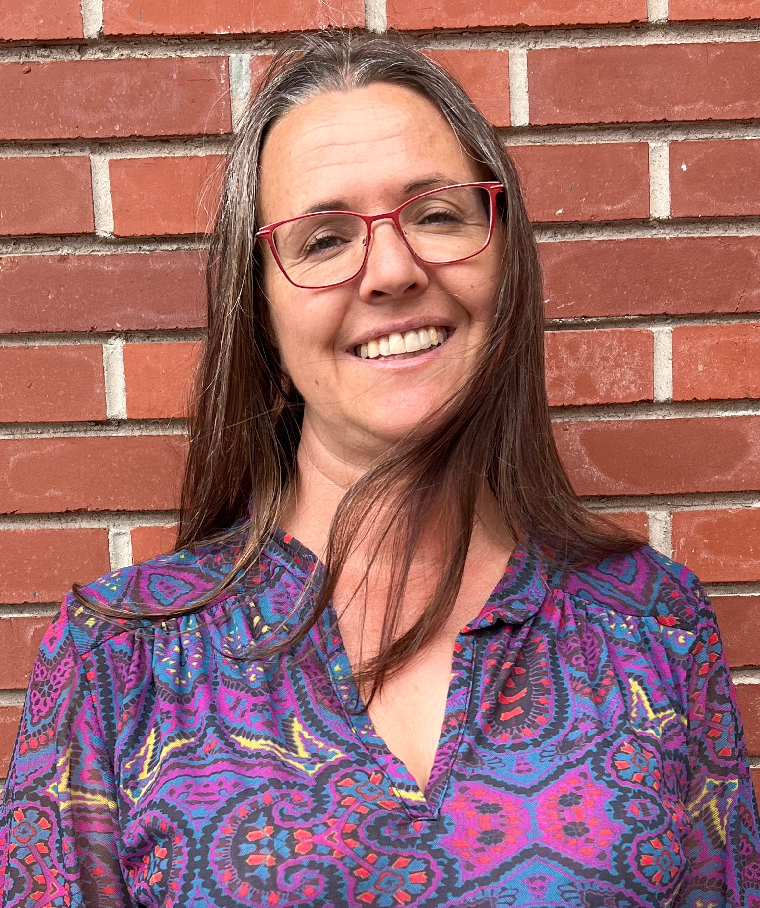Smiling woman with red glasses in a colorful patterned blouse stands against a red brick wall.