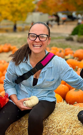 Smiling woman in glasses sits on hay, holding a small white pumpkin. Pumpkin patch background with autumn colors. Casual, joyful mood.