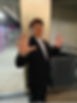 Man in a black suit stands indoors with hands raised, smiling. Concrete and metal background creates an industrial setting.