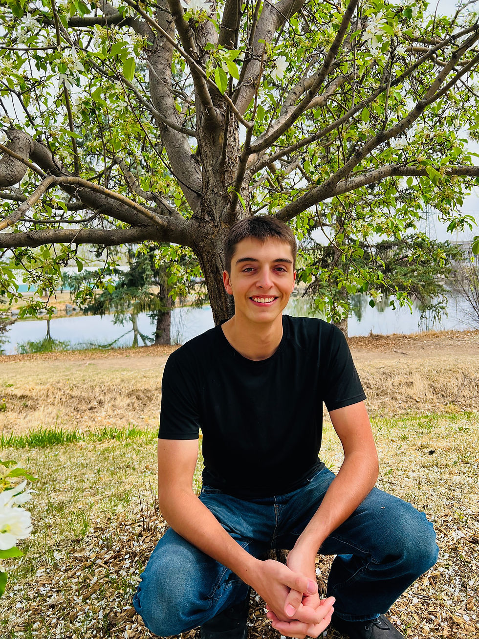 Young man in black t-shirt and jeans smiles while kneeling under a tree with green leaves. Background includes grass and distant water.