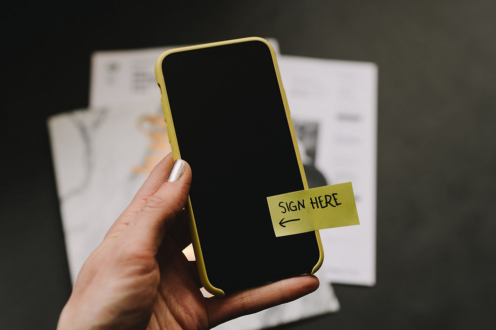 Kvenmanshönd heldur á snjallsíma með gulum bömper. Á símanum er gulur miði límdur neðarlega á svartan skjáinn. „Sign here" stendur á miðanum og ör bendir til vinstri í áttina að skjánum.