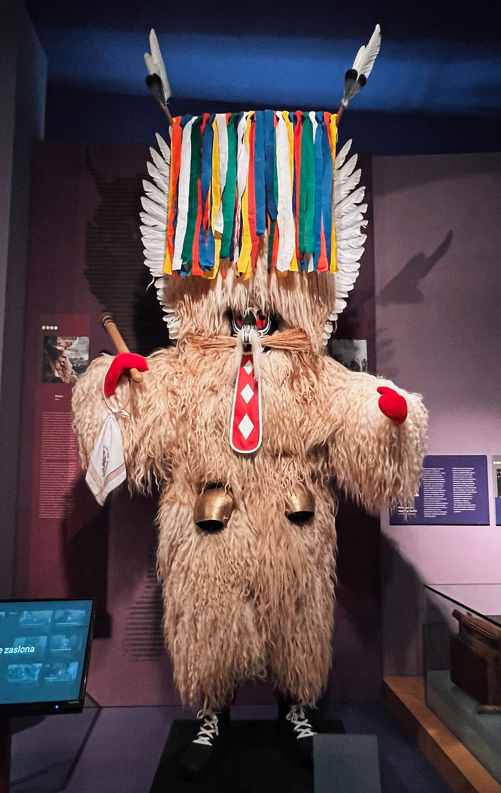 Furry costume with colorful ribbons and feathers, holding a staff and a cloth in a museum setting. Bells and patterns adorn the outfit.