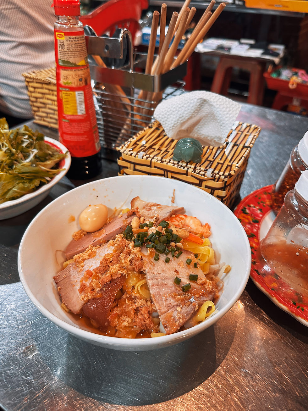 Bowl of noodles topped with pork, egg, and greens on a metal table. Red condiment bottle, chopsticks, and tissues in the background.