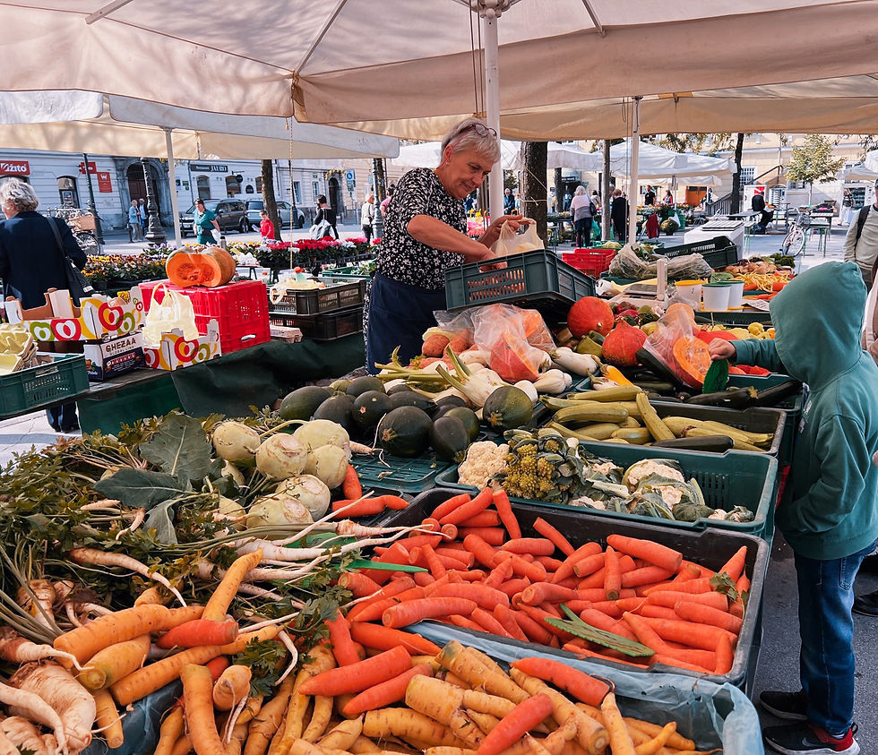 Outdoor market in Ljubljana with an elderly woman and a child selecting vegetables. Colorful produce and boxed goods under tents in a busy outdoor setting.