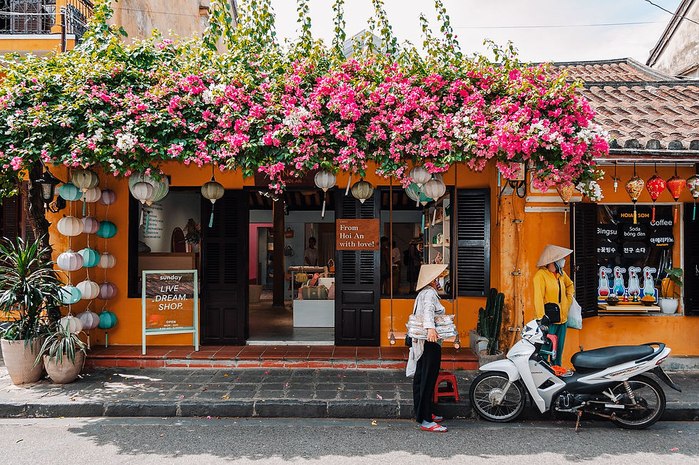 Street scene in Hoi An. Bright orange shop with pink flowers above. Two people wearing conical hats; one stands near a motorcycle. Sign: "From Hoi An with love!"