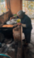 Elderly woman in a blue jacket stirs food in a rustic kitchen. Sunlit trees visible outside, creating a warm, serene atmosphere.