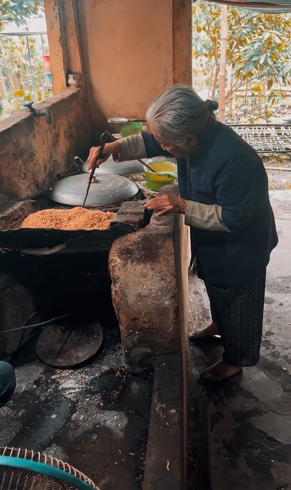 Elderly woman in a blue jacket stirs food in a rustic kitchen. Sunlit trees visible outside, creating a warm, serene atmosphere.
