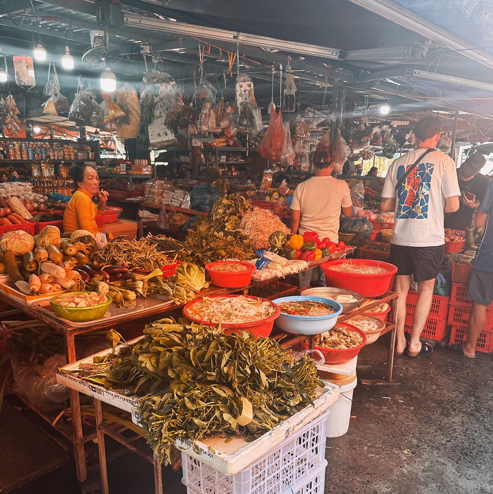 Vibrant market scene with people browsing stalls of vegetables and herbs in red bowls. Warm lighting creates a bustling, lively atmosphere.