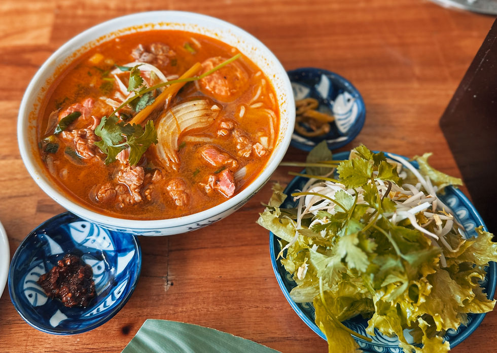 A bowl of spicy red soup with meat and herbs beside a plate of fresh greens and sprouts on a wooden table. Blue patterned dishes add detail.