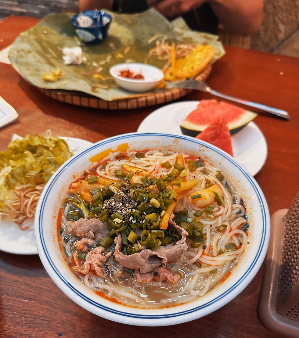 A bowl of noodle soup with vegetables and beef, beside a plate of greens and watermelon on a wooden table, evokes a cozy meal setting.