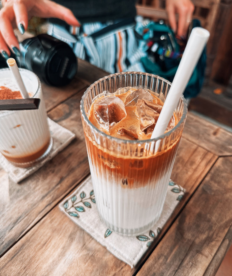 Iced coffee with milk in a ribbed glass, on a wooden table. A hand reaches for a camera. Striped clothing in the background. Relaxed vibe.