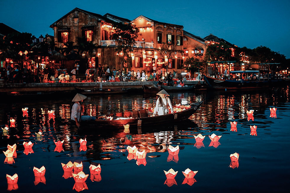 Two people in a boat on a lantern-lit river in the evening. Vibrant red and yellow lanterns float on water. Historic buildings glow in the background.