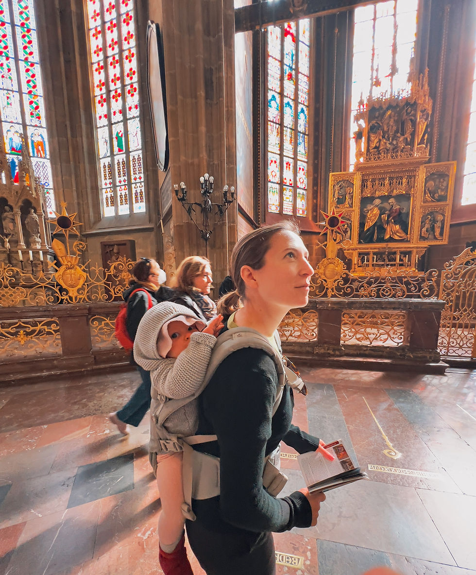 A woman carrying a baby in a church, gazing at stained glass in Prague. She holds a pamphlet. Warm light, ornate decor, and a peaceful ambiance.