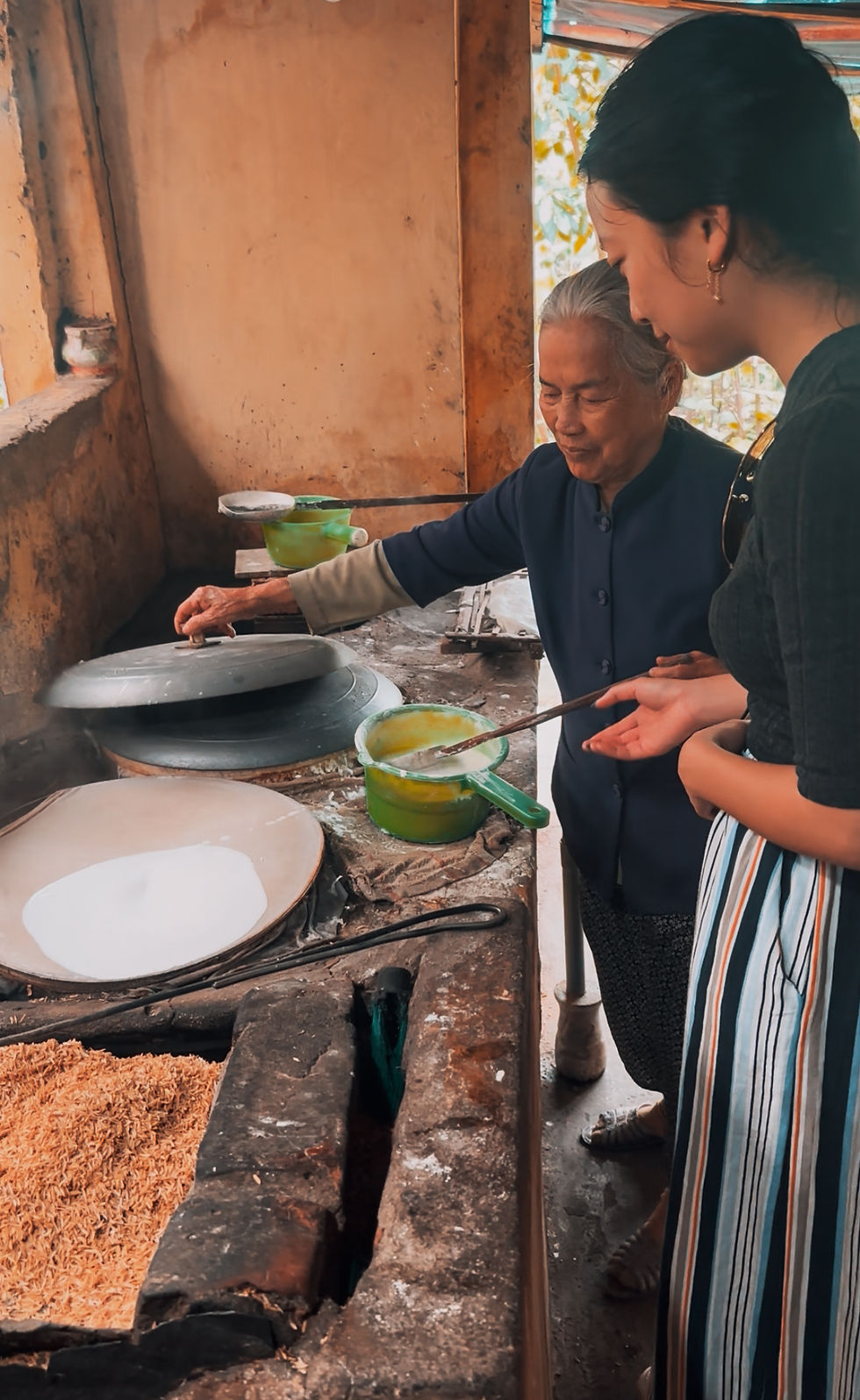 Elderly woman teaches young woman to cook over a rustic stove in a warm-toned kitchen, using a green pot and a ladle.