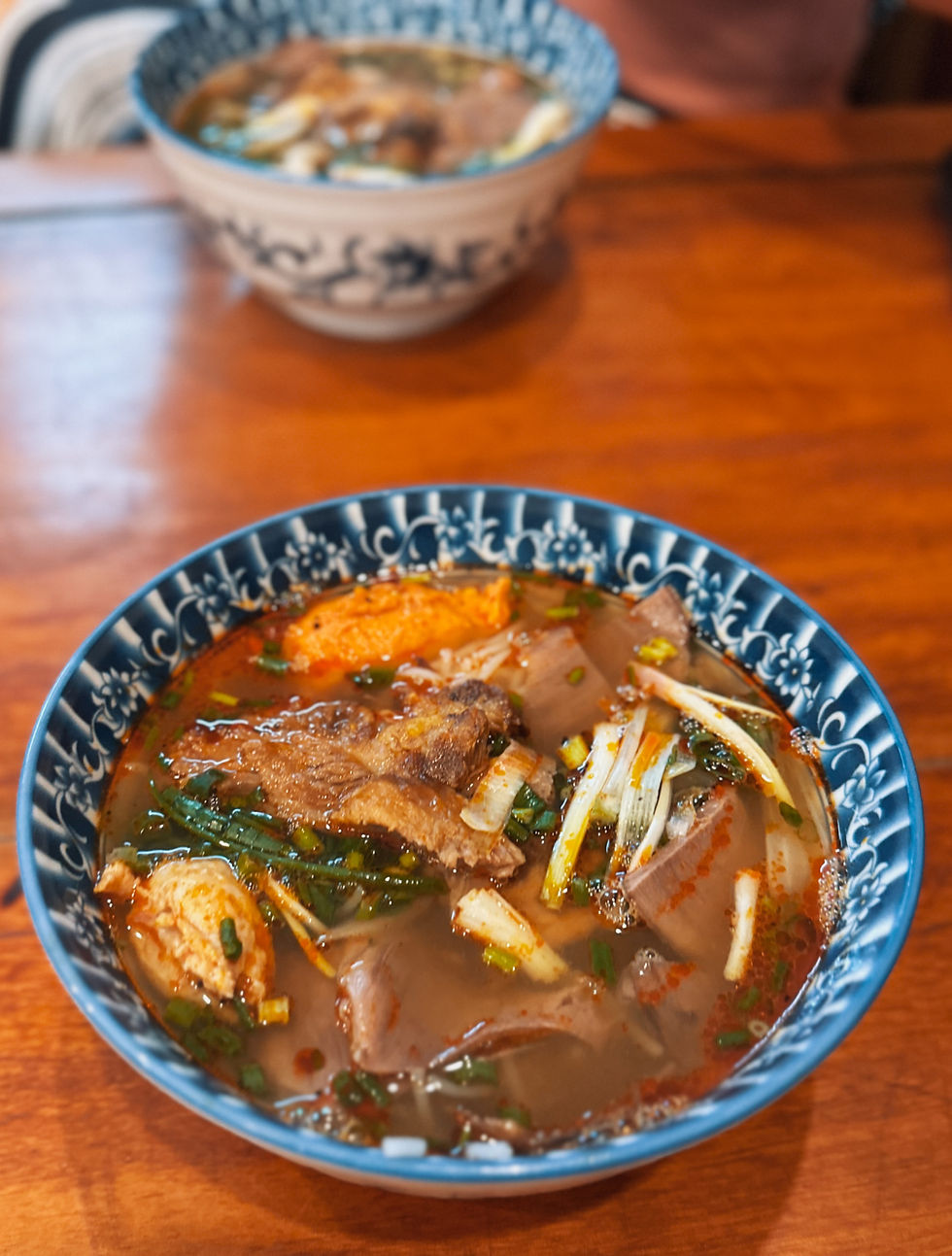 Blue bowl of noodle soup with meat, herbs, and spices on a wooden table. A similar bowl in the background. Warm, appetizing setting.