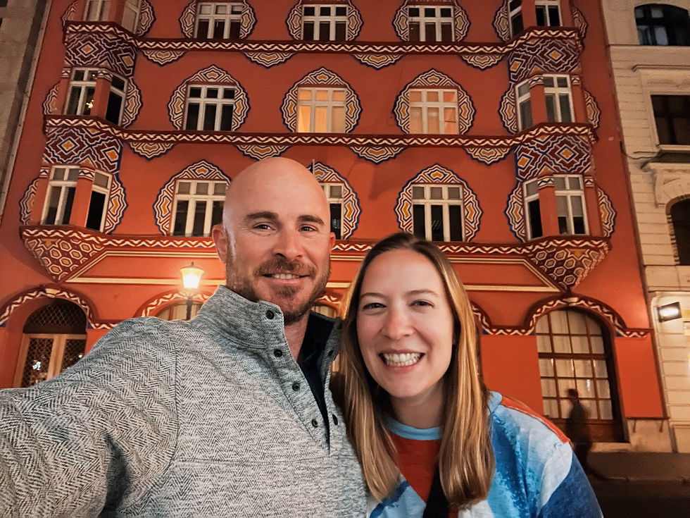 Smiling man and woman pose in front of a colorful, patterned building at night in Ljubljana. The structure has ornate designs and bright colors.