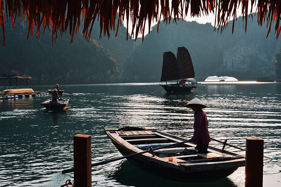 Boats glide in a serene bay as a person rows in the foreground wearing a conical hat. Lush hills and traditional sails are in the background.