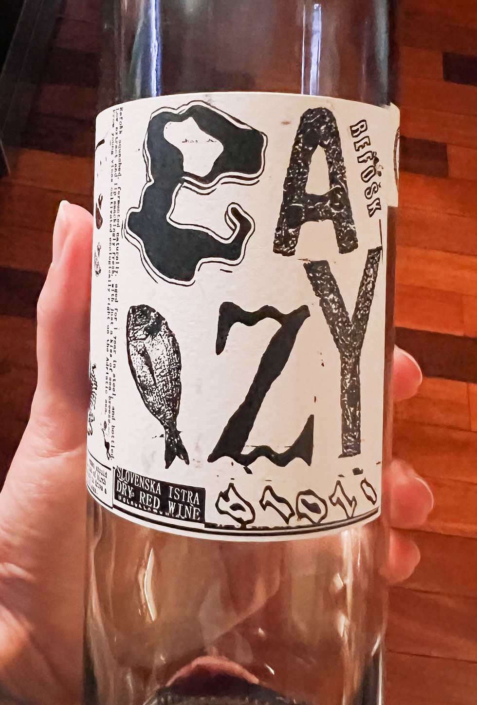 Hand holding a wine bottle with a label reading "EAZY" and "Slovenska Istra Dry Red Wine." Bold black text and abstract shapes. Wooden floor.