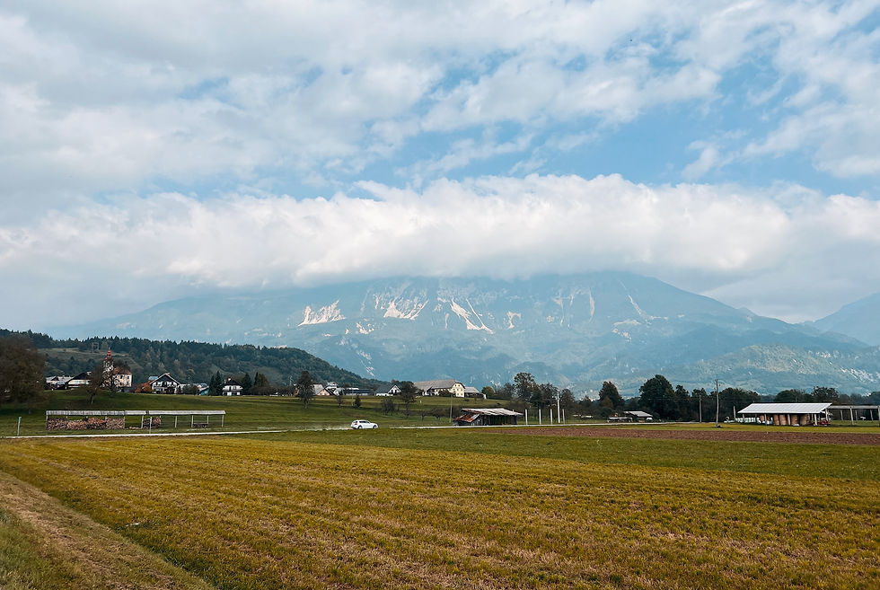 Vast green field with a white car driving on a rural road. Mountains shrouded in clouds form the backdrop. Blue sky and scattered clouds.