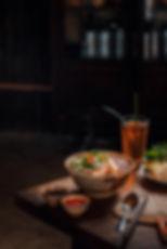 Steaming bowl of noodle soup with herbs on a wooden table, flanked by a tall iced tea, sauces, and garnishes, evoking a cozy, inviting mood.