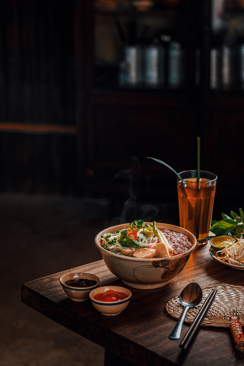 Steaming bowl of noodle soup with herbs on a wooden table, flanked by a tall iced tea, sauces, and garnishes, evoking a cozy, inviting mood.