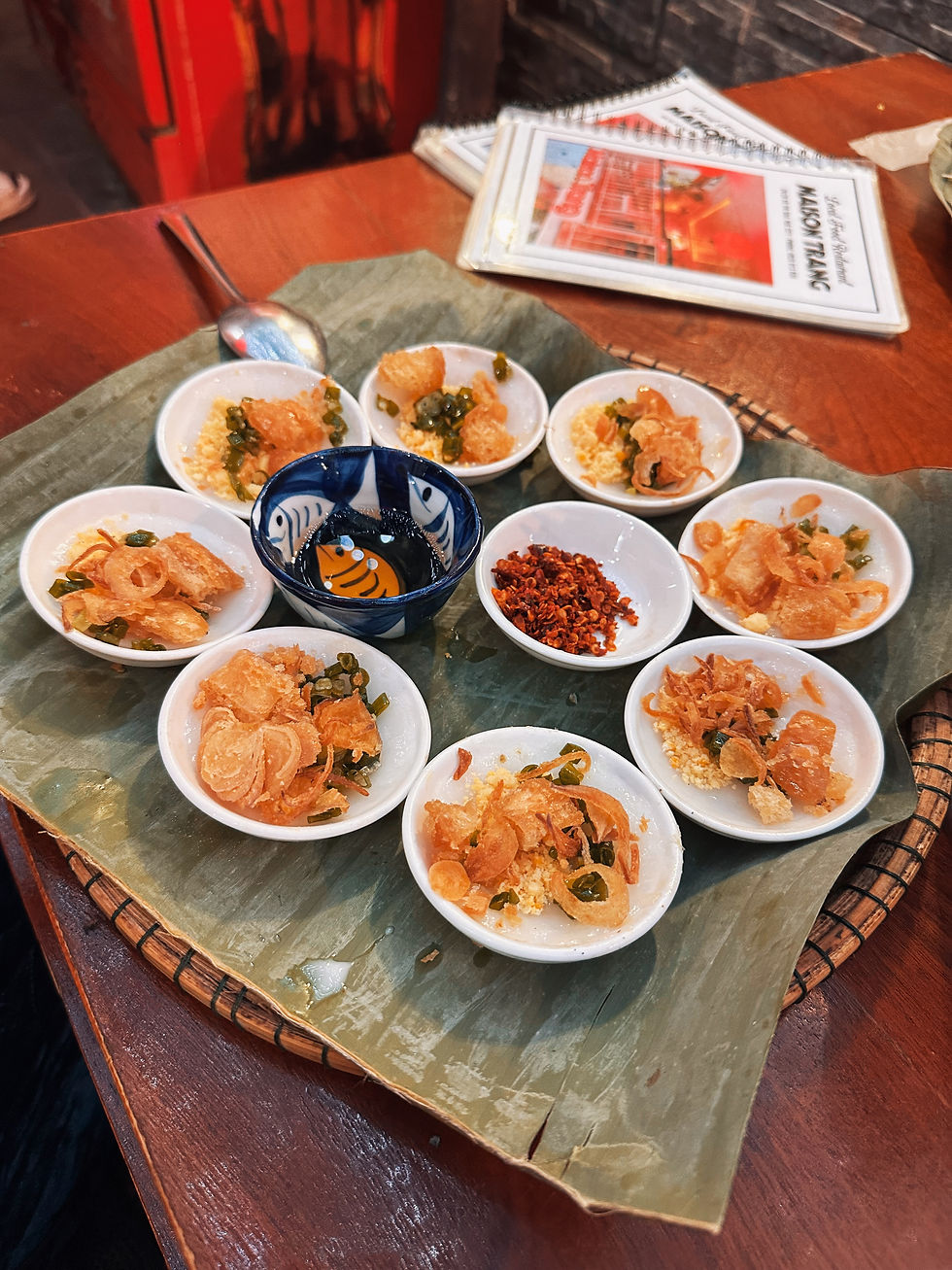 A platter of small bowls with fried toppings and chili on a banana leaf, surrounded by menus on a wooden table in a cozy setting.