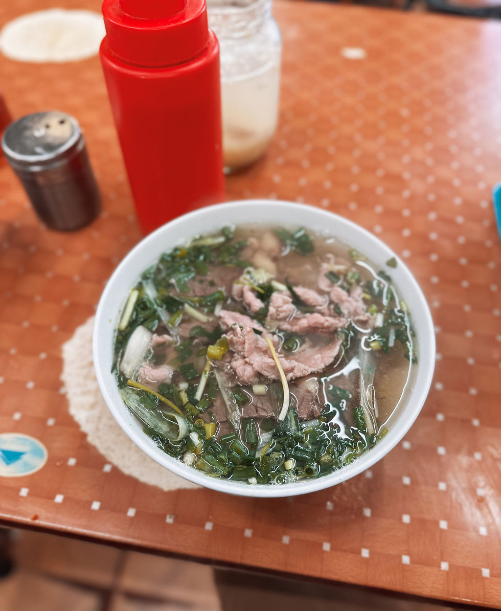 Bowl of soup with beef and greens on a patterned tablecloth. Red bottle and jar in the background create a cozy, inviting atmosphere.