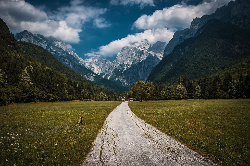 A winding path leads through a green meadow toward towering mountains under a cloudy blue sky, creating a serene, majestic landscape.