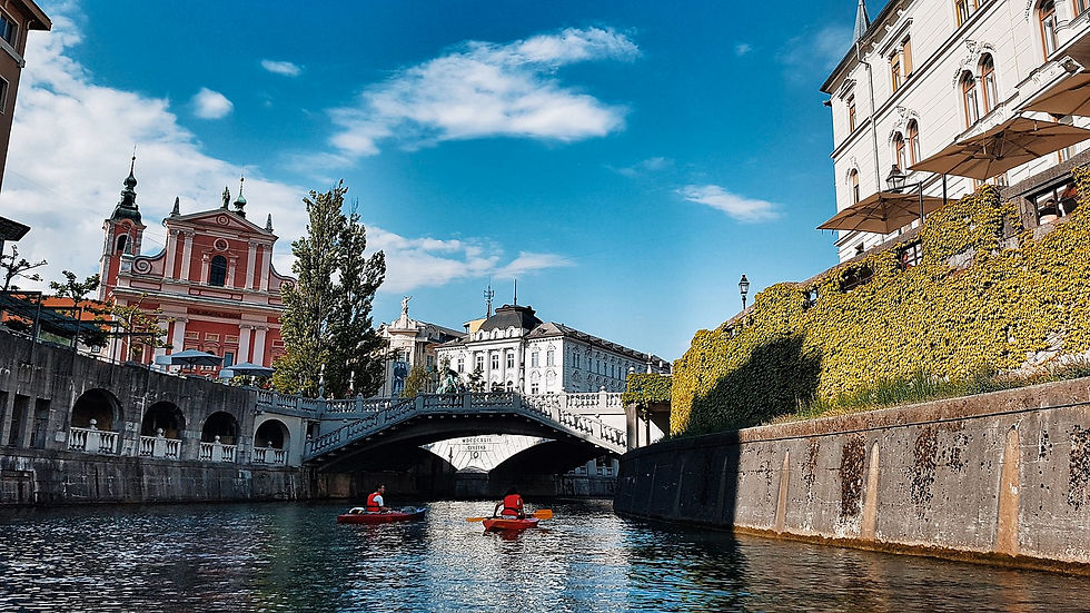 Two people in kayaks paddle under a bridge in a scenic canal in Ljubljana, Slovenia. A pink church and lush greenery adorn the vibrant urban landscape.