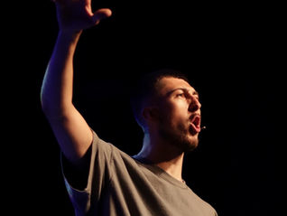 Facilitator David Woolcott lit up by a bright light on a dark stage. He has a small microphone attached to his ear, and is holding up one hand as if in the middle of explaining something.