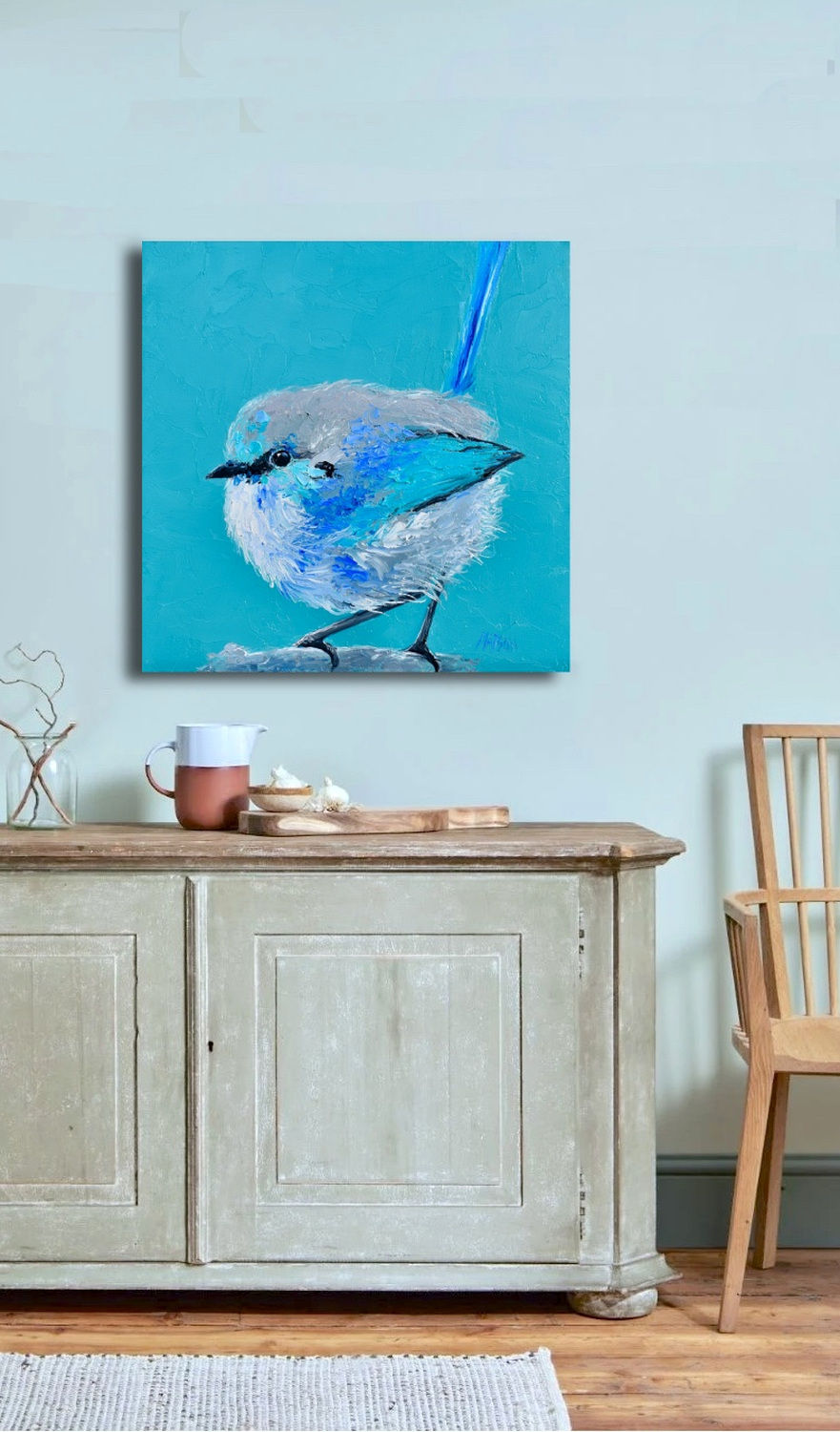 Room mockup of Blue Fairy Wren Bird Art on turquoise background by Jan Matson: the square turquoise fairy-wren painting hangs on a pale mint-blue wall above a rustic weathered wood sideboard styled with a glass vase of bare twigs, a two-tone terracotta-and-white ceramic pitcher, and a small wooden bowl on a cutting board; a light wood spindle-back chair sits to the right on timber floorboards with a woven rug at the edge — soft drop shadow under the canvas in a bright living-room setting.