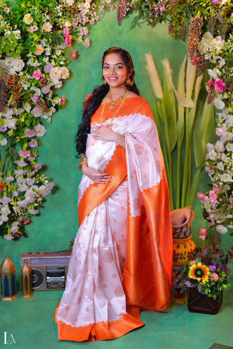 A smiling pregnant woman posing for a maternity photoshoot by Lensaction Photography, wearing a traditional white and orange silk saree against a floral green backdrop.