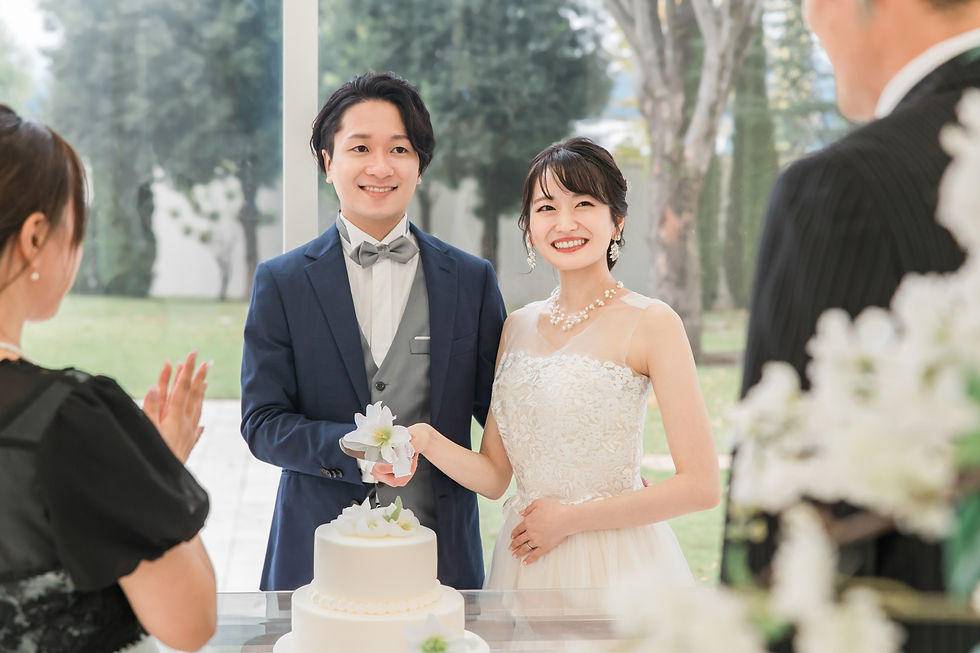 Bride and groom smiling while cutting a white wedding cake in a garden setting. Guests in formal attire clap, creating a joyful atmosphere.