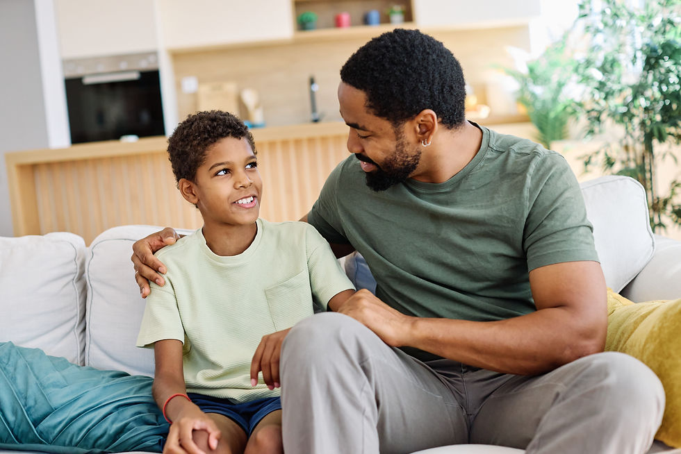 Father and son sit on a white couch, smiling at each other in a bright living room with warm, cozy decor.